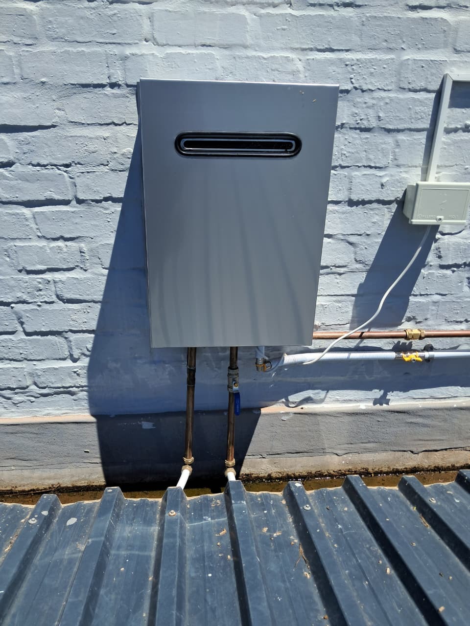 Grey gas geyser installation mounted on painted brick above a corrugated roof with copper pipework in George