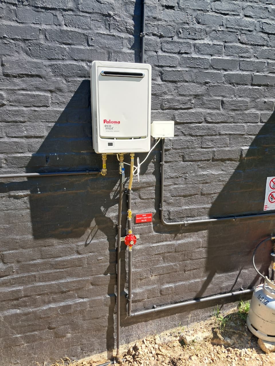 Paloma eco max gas geyser installation on a dark brick wall in George, with 19kg cylinder and red gas safety shut-off valve
